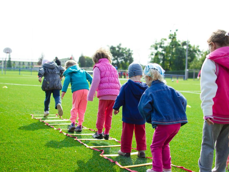 Children enjoy an outdoor activity on a grassy field, stepping over a ladder.