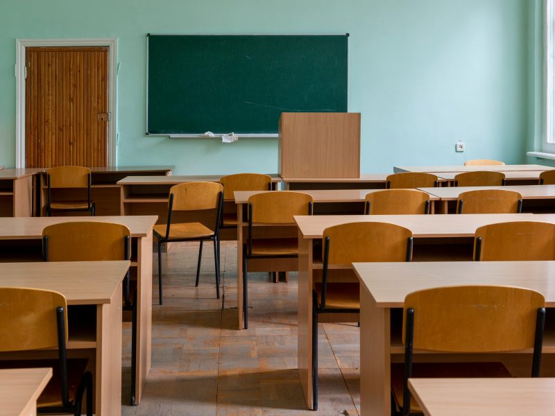 An empty classroom featuring wooden desks, chairs, and a large green chalkboard.