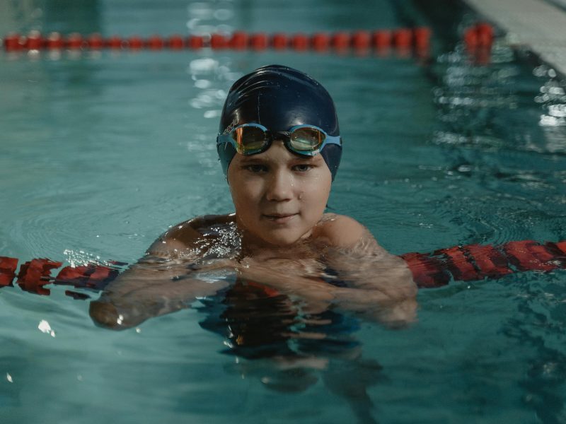 A young swimmer in a cap and goggles taking a break in an indoor pool.
