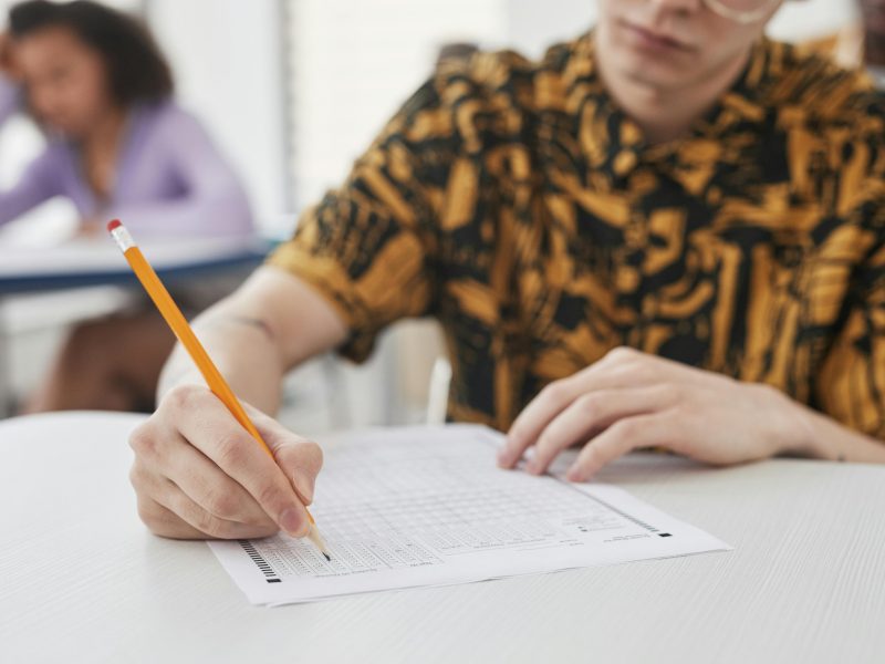Young adult writing on exam paper in classroom setting, focus on pencil and paper.