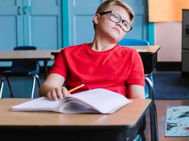 A young boy wearing glasses and a red shirt daydreams in an empty classroom, leaning back in his chair.