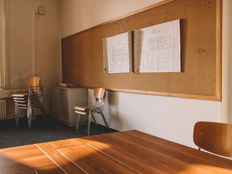 An empty classroom with stacked chairs, tables, and a cork board on the wall.